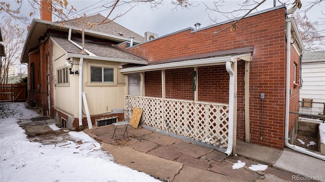 snow covered rear of property featuring roof with shingles, brick siding, a chimney, and fence