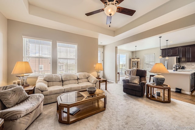 living room featuring a raised ceiling, baseboards, ceiling fan with notable chandelier, and light colored carpet