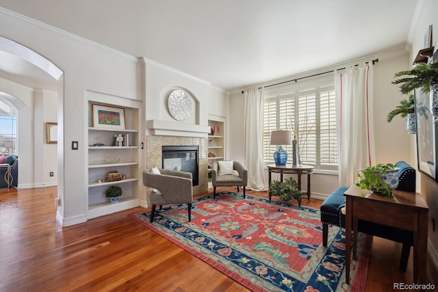 sitting room with arched walkways, wood finished floors, baseboards, a tiled fireplace, and crown molding