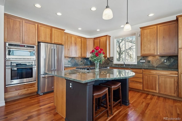 kitchen with a center island, pendant lighting, appliances with stainless steel finishes, brown cabinetry, and dark stone countertops