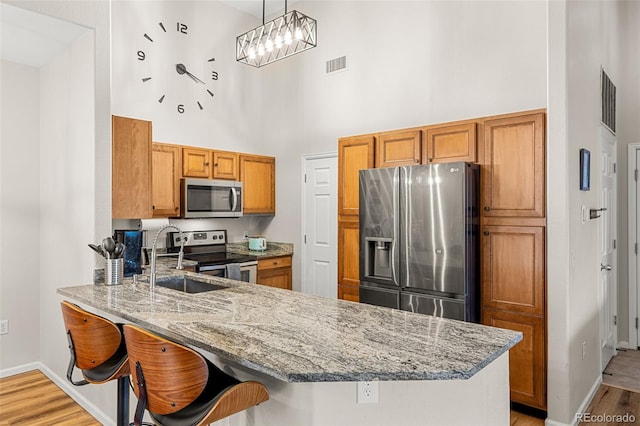 kitchen with brown cabinetry, a towering ceiling, appliances with stainless steel finishes, a sink, and a peninsula