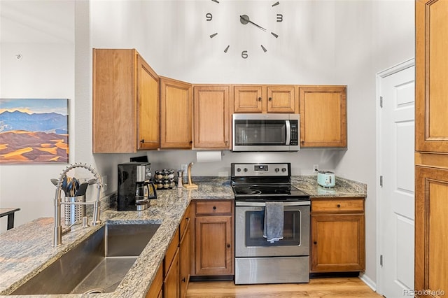 kitchen featuring appliances with stainless steel finishes, light stone counters, a sink, light wood finished floors, and brown cabinetry