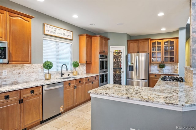 kitchen with backsplash, a sink, light stone counters, glass insert cabinets, and stainless steel appliances