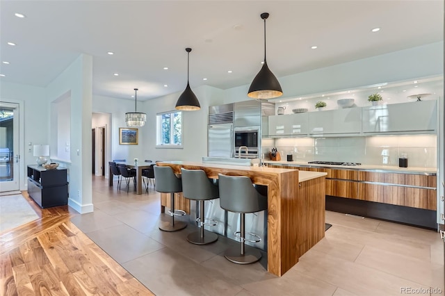 kitchen featuring decorative backsplash, built in appliances, a center island with sink, white cabinetry, and hanging light fixtures