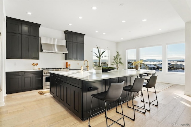 kitchen with a breakfast bar, light wood-type flooring, stainless steel range, a kitchen island with sink, and wall chimney range hood