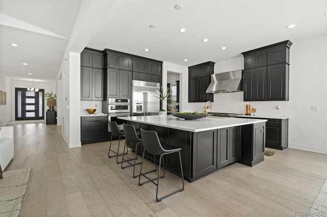 kitchen featuring wall chimney range hood, a breakfast bar area, appliances with stainless steel finishes, light hardwood / wood-style floors, and a large island with sink