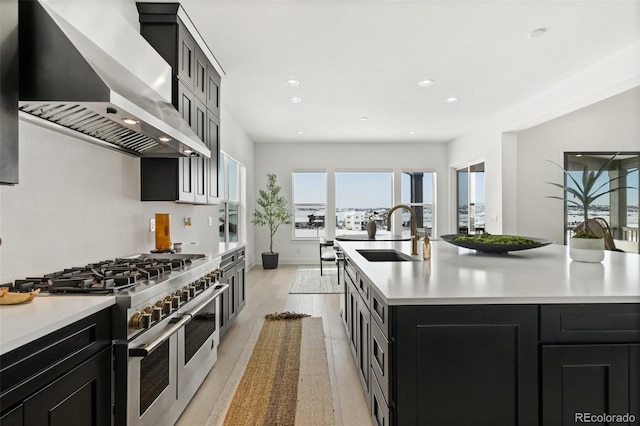 kitchen with wall chimney exhaust hood, sink, a center island with sink, light wood-type flooring, and range with two ovens