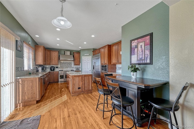 kitchen with high end appliances, wall chimney exhaust hood, brown cabinets, decorative light fixtures, and a center island