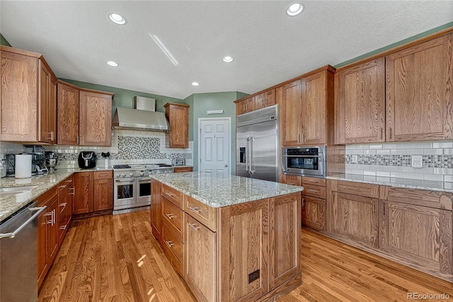 kitchen with light stone counters, brown cabinetry, a kitchen island, high quality appliances, and wall chimney exhaust hood