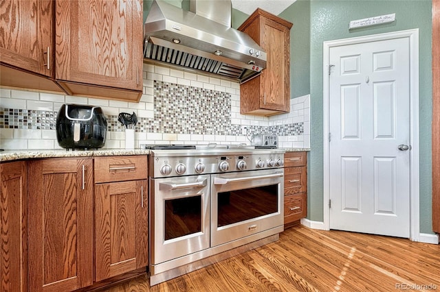 kitchen featuring range with two ovens, brown cabinets, light wood finished floors, tasteful backsplash, and wall chimney range hood