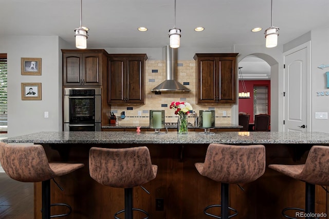 1315 W 141st Cir, Westminster, CO 80023 - kitchen featuring stainless steel double oven, wall chimney exhaust hood, arched walkways, a breakfast bar area, and dark brown cabinetry