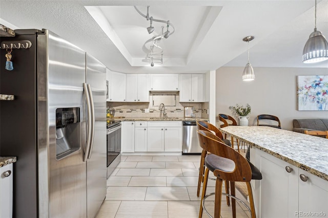 kitchen with light stone countertops, appliances with stainless steel finishes, a tray ceiling, white cabinets, and hanging light fixtures