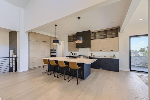 kitchen with light brown cabinetry, an island with sink, hanging light fixtures, light hardwood / wood-style floors, and wall chimney exhaust hood