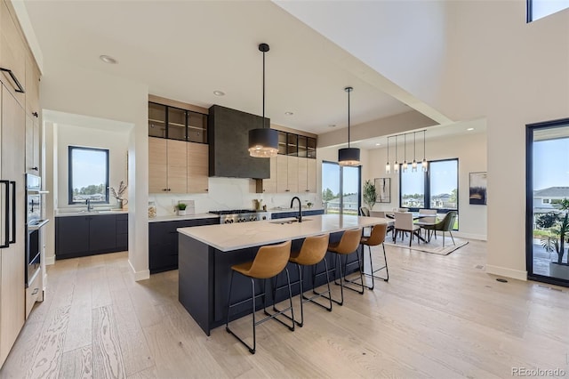 kitchen with an island with sink, wall chimney range hood, light brown cabinets, and decorative light fixtures