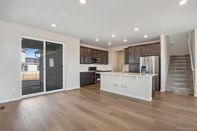 kitchen featuring dark brown cabinetry, appliances with stainless steel finishes, a kitchen island with sink, light countertops, and light wood-style floors