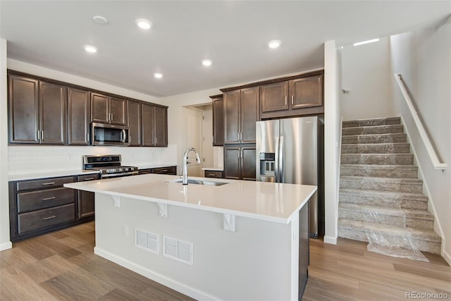 kitchen with light wood finished floors, appliances with stainless steel finishes, a kitchen island with sink, light countertops, and a sink