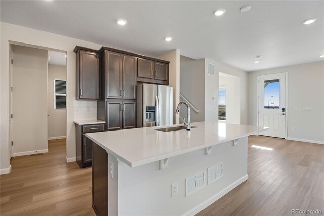 kitchen featuring a center island with sink, light countertops, a sink, dark brown cabinets, and stainless steel fridge