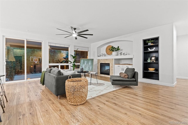 living room with built in shelves, ceiling fan, light wood-type flooring, and a tiled fireplace