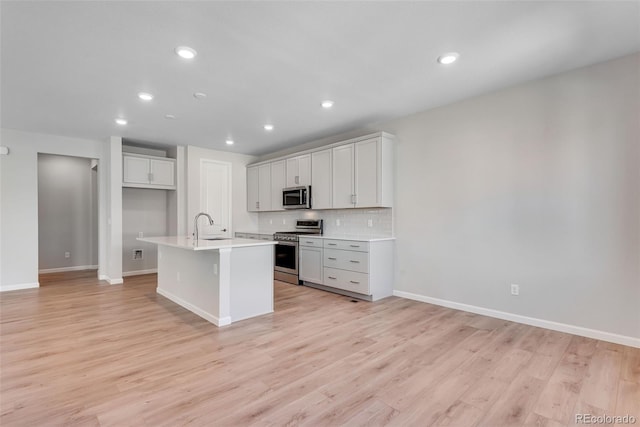 kitchen with a sink, light wood-type flooring, an island with sink, backsplash, and appliances with stainless steel finishes