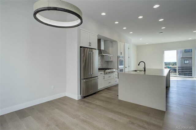 kitchen featuring stainless steel appliances, wall chimney exhaust hood, sink, a center island with sink, and light hardwood / wood-style flooring