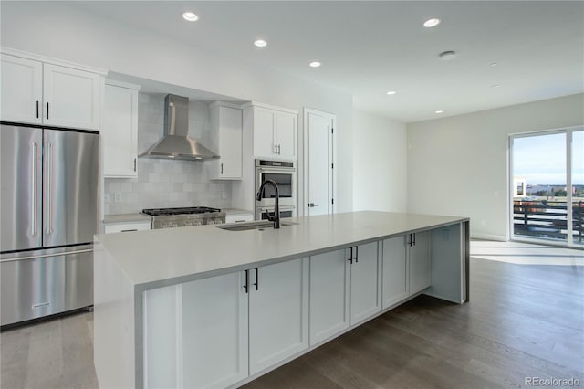 kitchen featuring tasteful backsplash, an island with sink, hardwood / wood-style floors, wall chimney exhaust hood, and appliances with stainless steel finishes