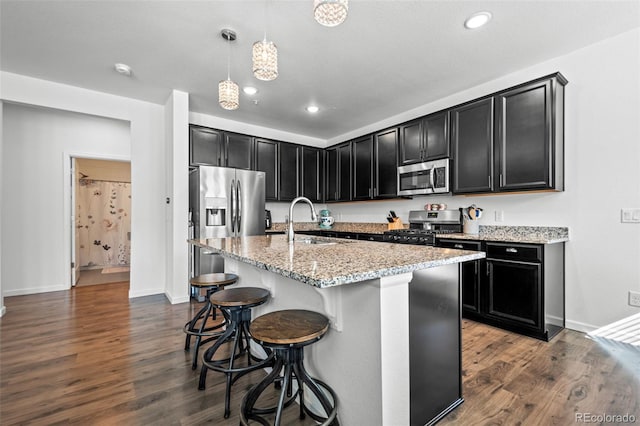 kitchen with light stone counters, pendant lighting, stainless steel appliances, a kitchen island with sink, and a sink