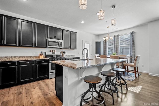 kitchen with stainless steel appliances, a sink, dark cabinetry, a center island with sink, and pendant lighting