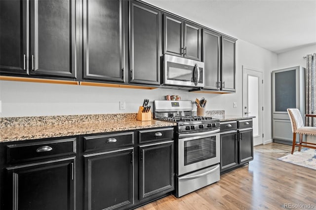 kitchen with appliances with stainless steel finishes, dark cabinetry, light stone counters, and light wood finished floors