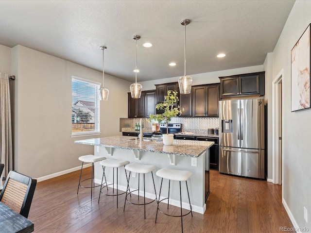 kitchen with pendant lighting, light stone countertops, a kitchen island with sink, decorative backsplash, and stainless steel appliances