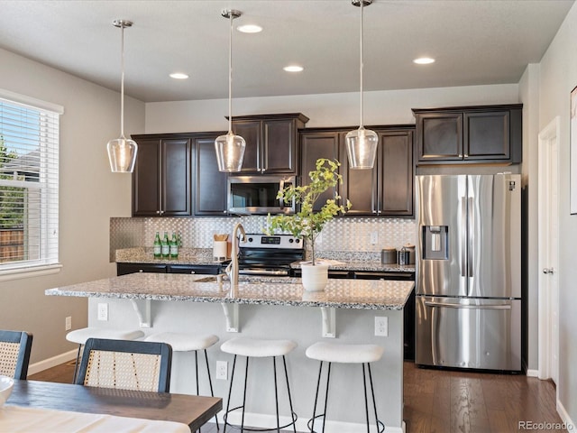 kitchen featuring light stone countertops, dark brown cabinetry, stainless steel appliances, an island with sink, and hanging light fixtures