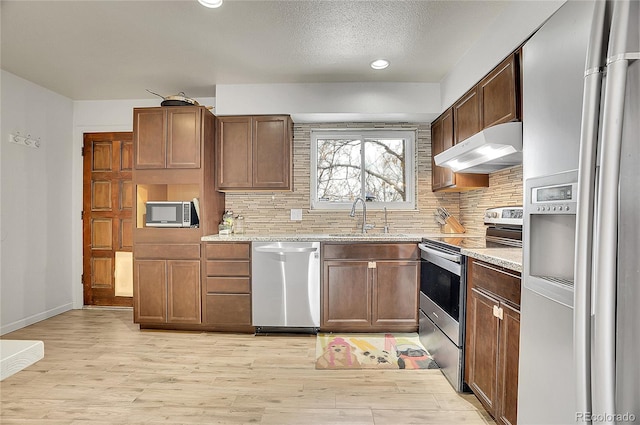 kitchen with light wood finished floors, stainless steel appliances, backsplash, a sink, and under cabinet range hood