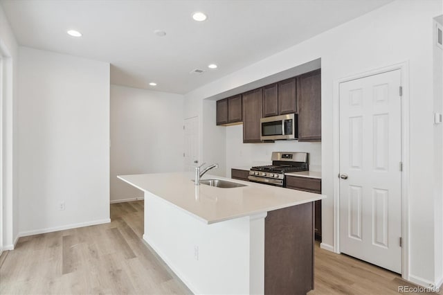 kitchen featuring sink, an island with sink, light hardwood / wood-style floors, dark brown cabinets, and appliances with stainless steel finishes