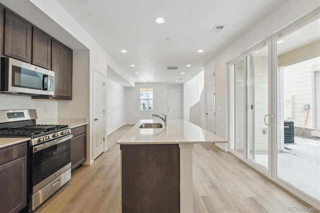 kitchen with dark brown cabinets, stainless steel appliances, a kitchen island with sink, sink, and light hardwood / wood-style flooring