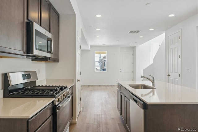 kitchen with dark brown cabinets, stainless steel appliances, a kitchen island with sink, sink, and light hardwood / wood-style flooring