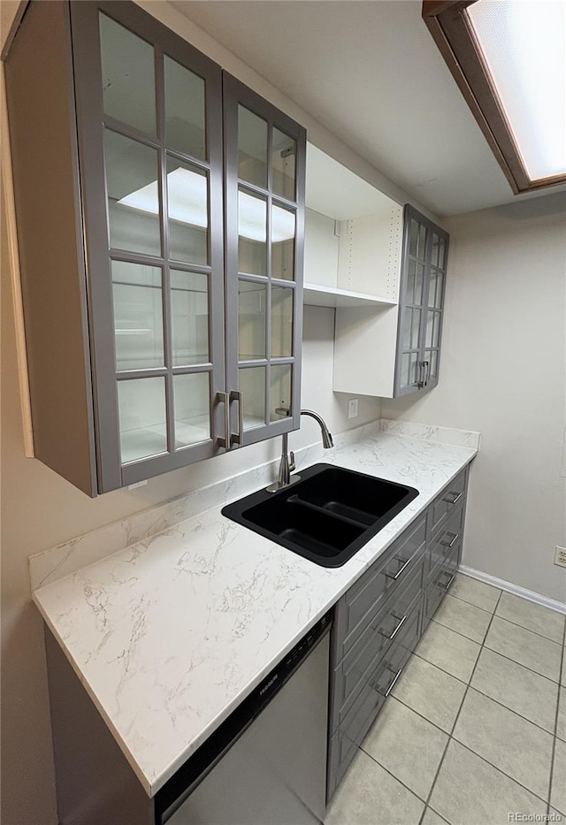 kitchen with light tile patterned floors, light stone counters, a sink, stainless steel dishwasher, and glass insert cabinets