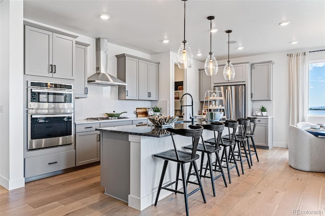 kitchen with wall chimney exhaust hood, an island with sink, light hardwood / wood-style floors, a breakfast bar, and appliances with stainless steel finishes