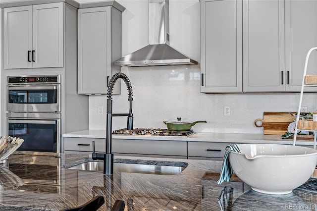 kitchen with double oven, gray cabinetry, dark stone countertops, and wall chimney range hood