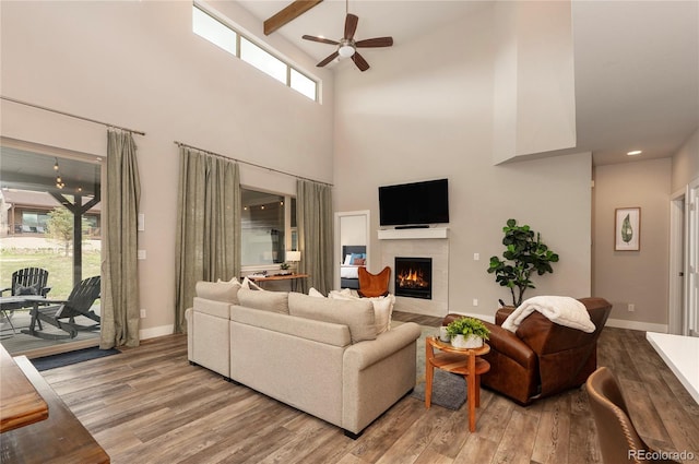 living room with ceiling fan, beamed ceiling, a high ceiling, a fireplace, and light hardwood / wood-style floors