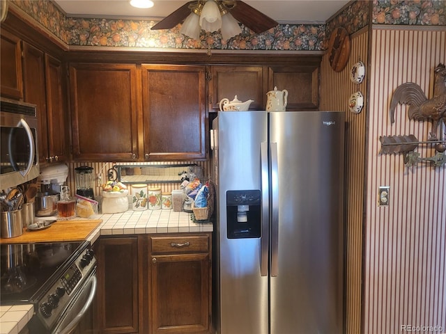 kitchen with tile countertops, appliances with stainless steel finishes, a ceiling fan, and wallpapered walls