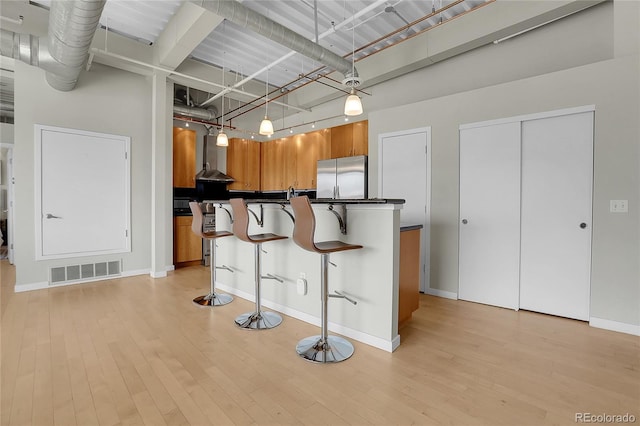 kitchen featuring hanging light fixtures, wall chimney range hood, stainless steel refrigerator, light wood-type flooring, and a kitchen bar