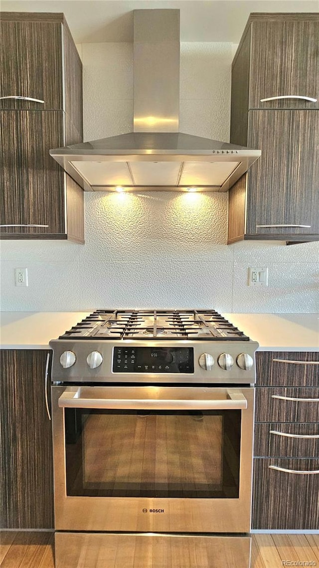 kitchen featuring stainless steel gas range oven, wall chimney range hood, backsplash, and dark brown cabinetry