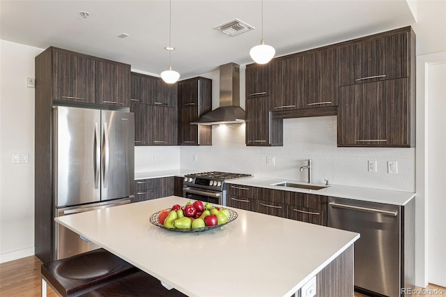 kitchen with sink, hanging light fixtures, stainless steel appliances, a kitchen island, and wall chimney exhaust hood