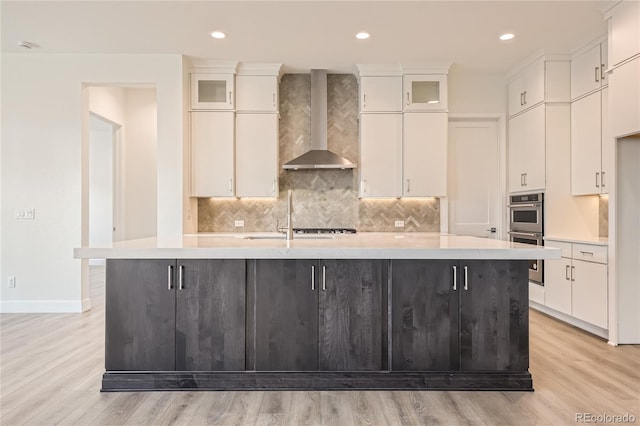 kitchen with white cabinets, light wood-type flooring, an island with sink, and wall chimney range hood