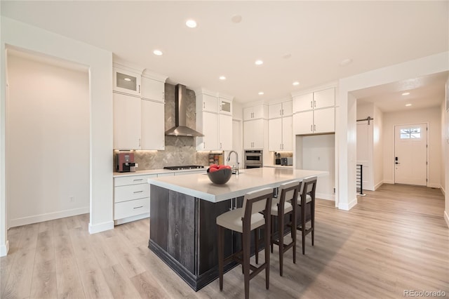 kitchen featuring a kitchen breakfast bar, wall chimney exhaust hood, a kitchen island with sink, light hardwood / wood-style flooring, and white cabinets