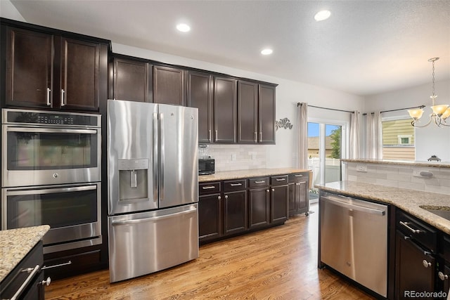 kitchen featuring stainless steel appliances, a chandelier, tasteful backsplash, dark brown cabinetry, and light stone countertops