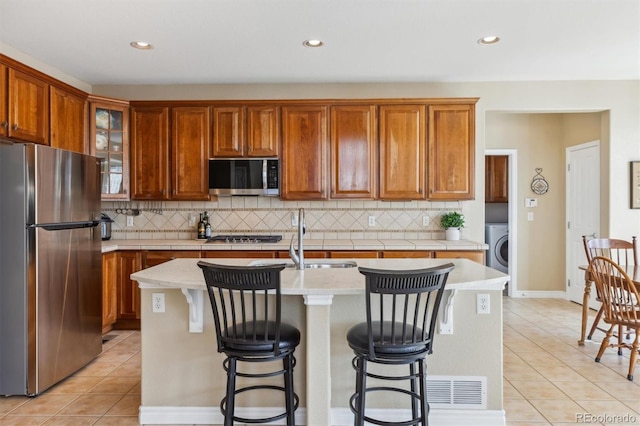 kitchen featuring light countertops, washer / dryer, appliances with stainless steel finishes, light tile patterned flooring, and a kitchen island with sink