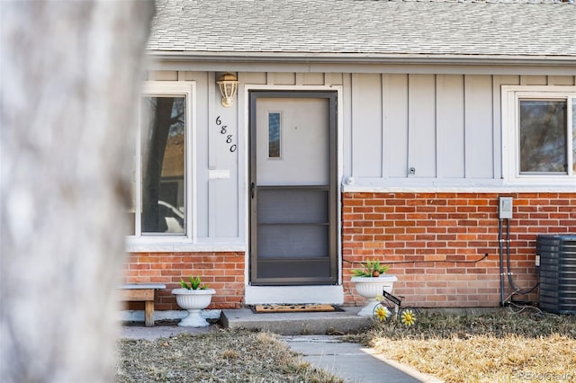 view of exterior entry with board and batten siding, brick siding, roof with shingles, and cooling unit