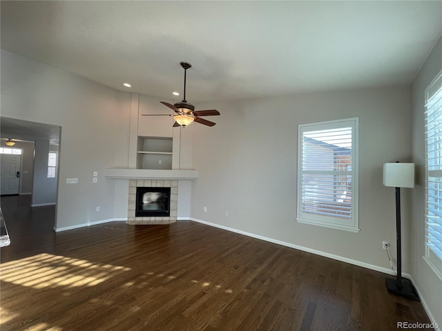 unfurnished living room with dark wood-type flooring, a tiled fireplace, a ceiling fan, and baseboards