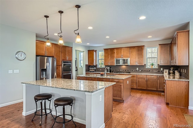 kitchen with light stone counters, stainless steel appliances, a kitchen island with sink, dark wood-type flooring, and decorative light fixtures