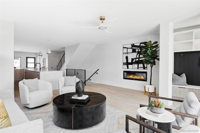 living area featuring light wood-style flooring, a ceiling fan, a glass covered fireplace, baseboards, and stairs